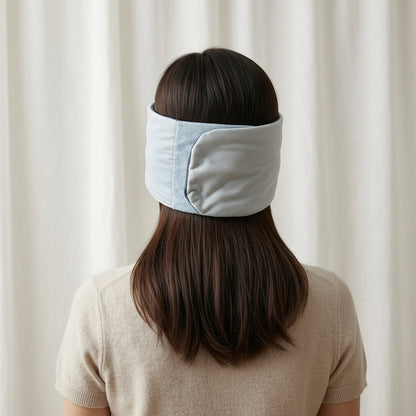 A woman viewed from behind wearing a SomniGrave Weighted Sleep Mask with cooling ice silk side with secure adjustable fit, against a soft cream curtain background.